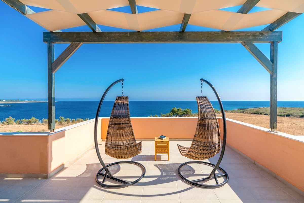 A balcony with two wicker chairs and a small table, overlooking a coastal view.