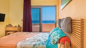 Ein Schlafzimmer mit Bett, Schreibtisch, Stuhl, Fernseher und einem Balkon mit Meerblick. An der Wand hängt ein Gemälde.