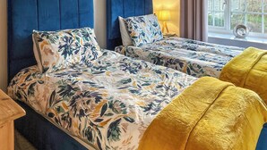 A double bed with floral bedding and a blue headboard, a bedside table with a lamp, and a view of the outdoors through a window.