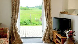 A room with a television, a wooden cabinet, and a view of a green field through curtains. - 2 bedroom accommodation in South Barrow, near Yeovil (South Barrow, near Yeovil)