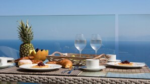 Morgenmad med ananas, croissanter og vinglas på en balkon med udsigt over havet.