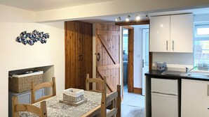 Een kleine keuken met houten kastjes, een eettafel met stoelen, en uitzicht op de aangrenzende badkamer.