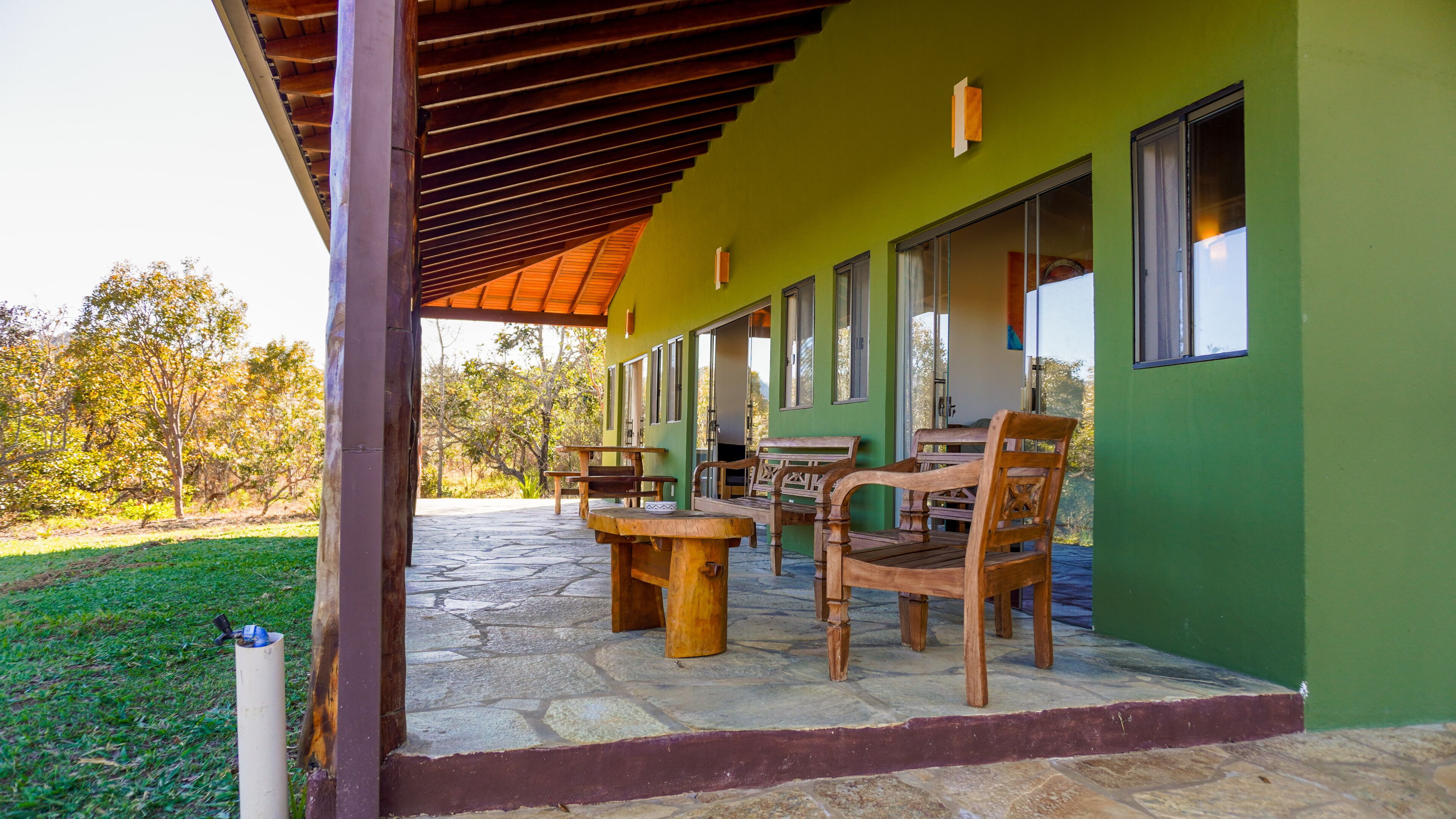Un patio exterior con suelo de piedra, muebles de madera y un edificio verde con ventanas amplias.