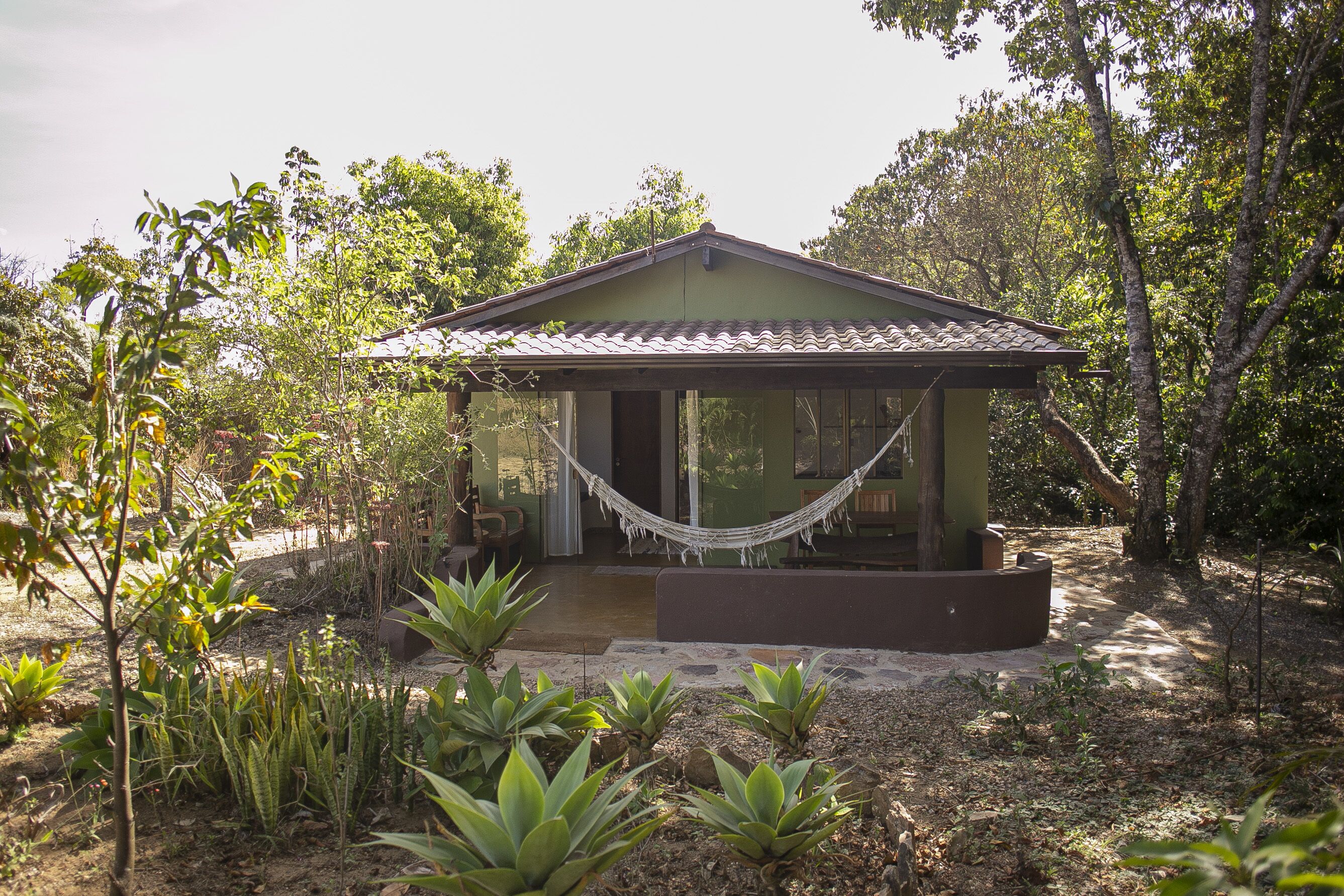 A small, single-story house with a tiled roof, surrounded by lush greenery and a hammock.