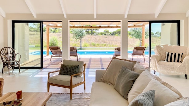 Un salon moderne avec un grand canapé, un fauteuil et une table à manger. On peut apercevoir une piscine à travers les portes vitrées.