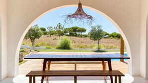 Une table et un banc en bois, placés sous un luminaire suspendu, avec vue sur une piscine et des arbres à l’extérieur.