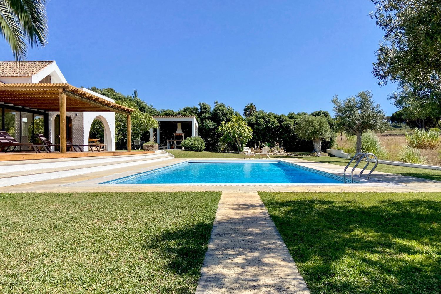 A swimming pool with a wooden deck and lounge chairs, surrounded by a well-maintained lawn and trees.