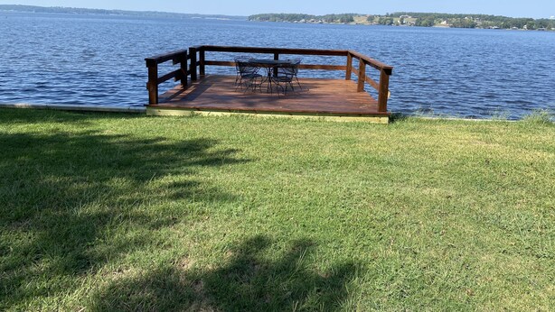 Une terrasse en bois avec une table et des chaises en métal, donnant sur une étendue d’eau.