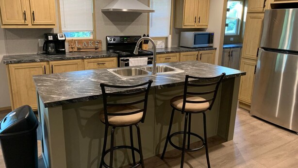 A kitchen with wooden cabinets, a stainless steel refrigerator, a microwave, a stove, and a sink. There are two bar stools with cushioned seats.