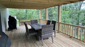 A covered outdoor deck with a table and chairs, surrounded by a wooden railing and overlooking a forested area.