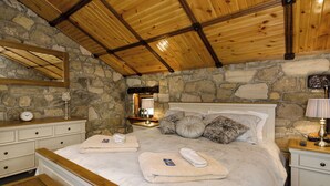 Une chambre avec un mur en pierre, des poutres en bois au plafond, un lit avec des oreillers et une couverture, une table de chevet avec une lampe et une petite table de nuit avec une horloge.