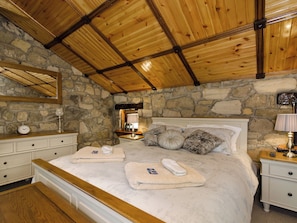 Une chambre avec un mur en pierre, des poutres en bois au plafond, un lit avec des oreillers et une couverture, une table de chevet avec une lampe et une petite table de nuit avec une horloge.