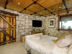 Een slaapkamer met een stenen muur, houten balken in het plafond, een bed met bloemenbeddengoed, een televisie en een raam met uitzicht.