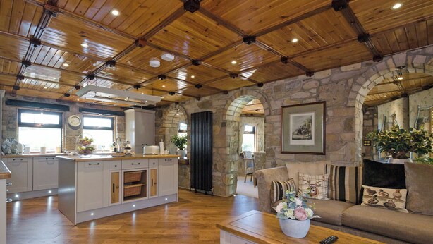Un vaste espace de vie doté d’un plafond en bois, de murs en pierre, d’un îlot de cuisine et d’un canapé agrémenté de coussins décoratifs.