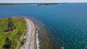 An aerial view of a coastal area with a rocky shoreline, clear blue water, and a distant tree line.