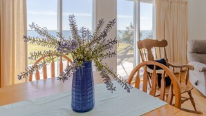 A dining area with a table, chairs, a vase with flowers, and large windows with a view of the outdoors.