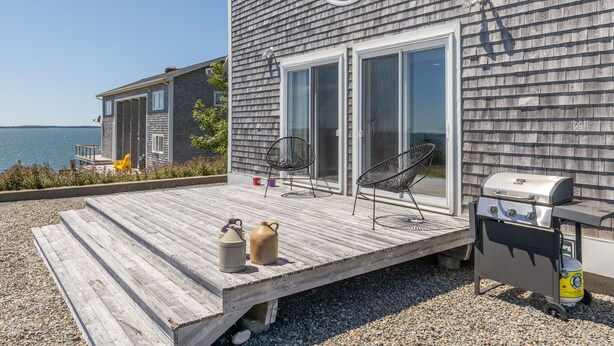 A wooden deck with a grill and outdoor chairs, overlooking a body of water.