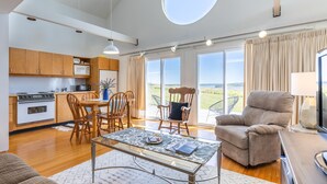 A modern kitchen with wooden cabinets, a dining area with a table and chairs, a sofa, and a large window offering a view.