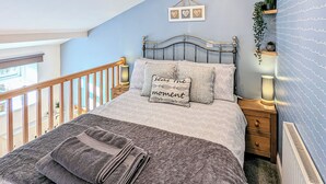 A bedroom with a bed, pillows, a bedside table, a nightstand, a wall-mounted shelf with plants, and a view of the outside. - The Hideaway Low Hawsker (Low Hawsker, near Whitby)