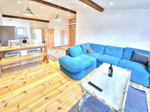 A modern living room with a blue sectional sofa, wooden flooring, a dining area with a wooden table and chairs, and a kitchen in the background. - 2 bedroom accommodation in Wellow, near Yarmouth (Wellow, near Yarmouth)