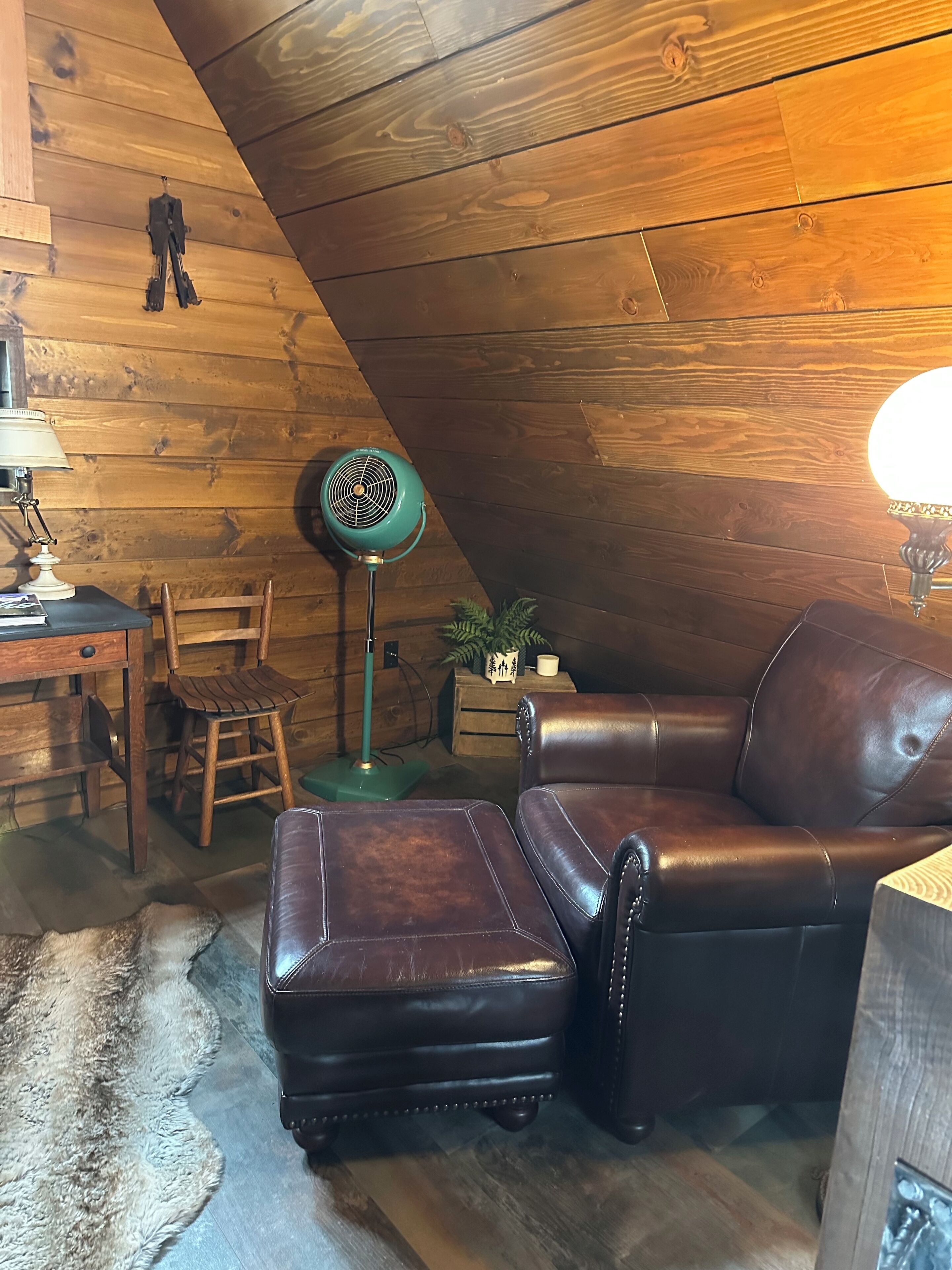 Um quarto aconchegante com um sofá de couro marrom, um banquinho combinando, uma escrivaninha de madeira com uma luminária, um abajur verde e uma pequena planta em vaso.