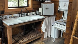 A rustic kitchen with wooden walls, a large window, a sink, a stove, and a microwave. - Cabin in the Meadow (Sandpoint)