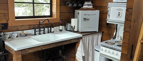A rustic kitchen with wooden walls, a large window, a sink, a stove, and a microwave.