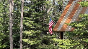 A deer standing in a garden area with a rustic building and a flag in the background. - Cabin in the Meadow (Sandpoint)