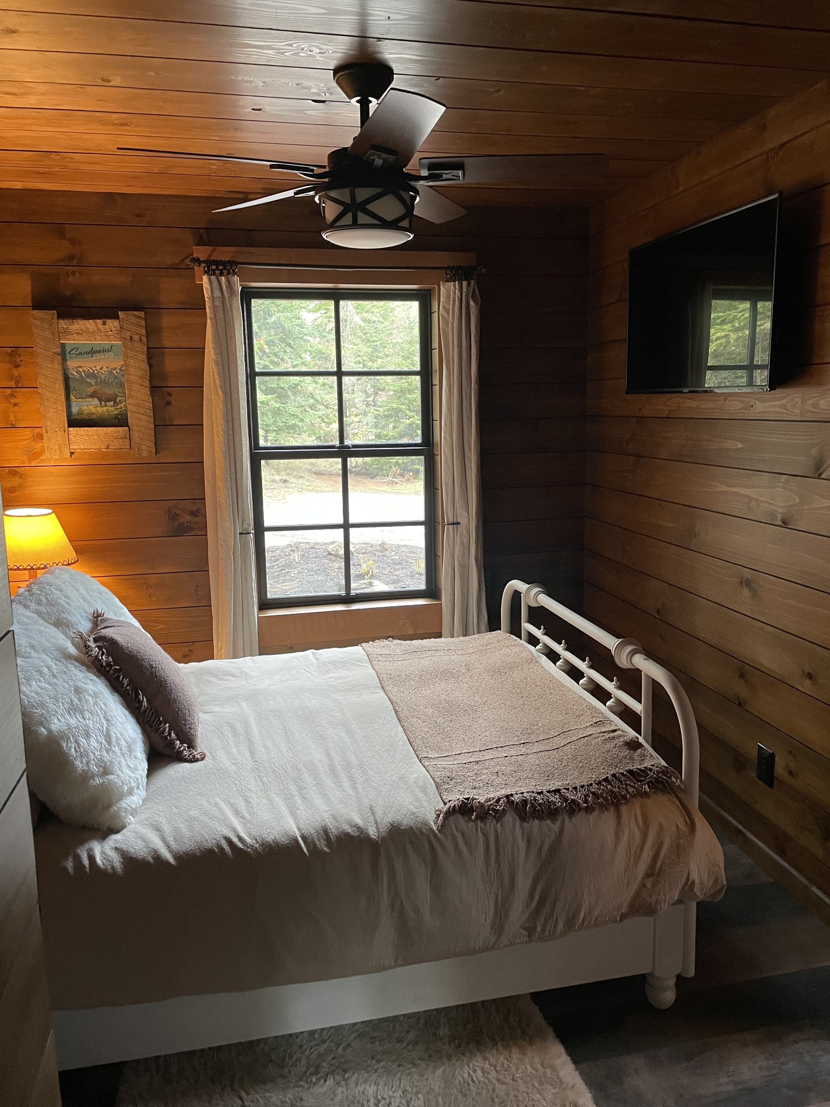 Un dormitorio de cabaña de madera con cama, ventilador de techo, ventana y lámpara.