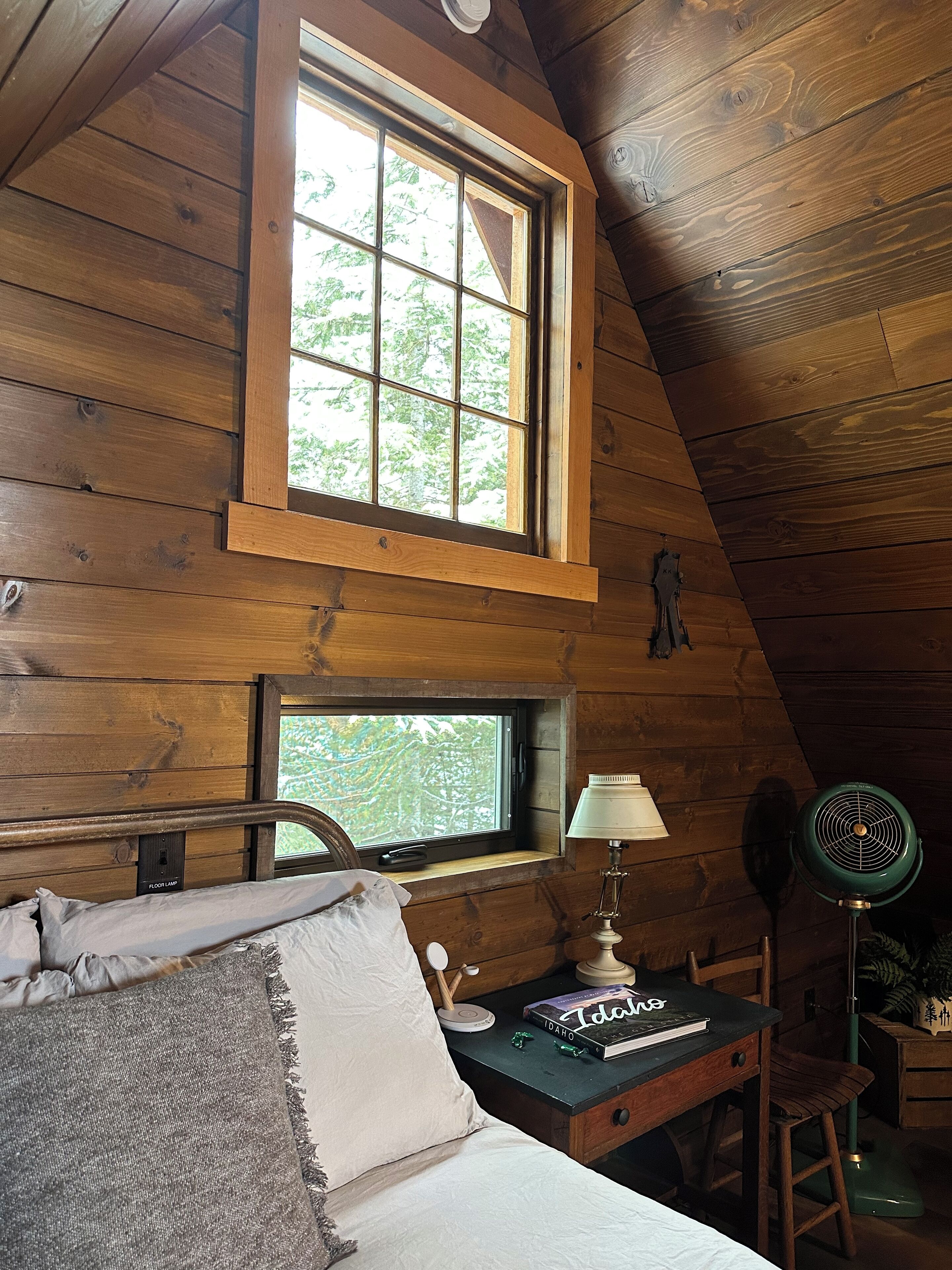 A wooden cabin room with a bed, a desk with a lamp, a chair, and a window with a view of trees.
