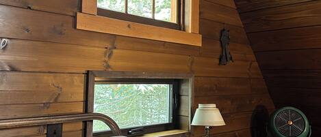 Une chambre dans un chalet en bois, avec un lit, un bureau doté d’une lampe, une chaise et une fenêtre donnant sur la forêt.