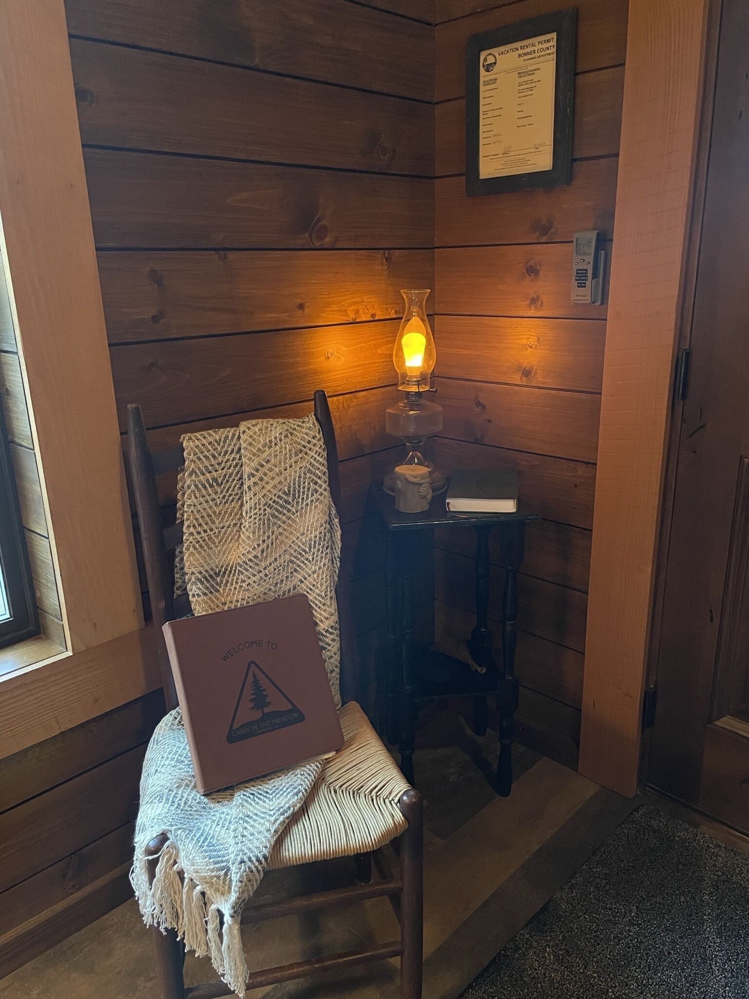 A wooden cabin interior with a chair holding a welcome box, a lit table lamp, and a framed rental permit on the wall.