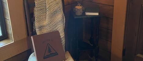 A wooden cabin interior with a chair holding a welcome box, a lit table lamp, and a framed rental permit on the wall.