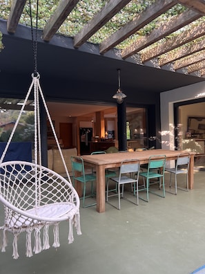 A wooden dining table with six chairs, a hanging macramé chair, and a hanging light fixture in a covered outdoor area.