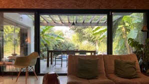 Un salón moderno con sofá, sillas y mesa de comedor. Amplias ventanas ofrecen vistas a un jardín exuberante.