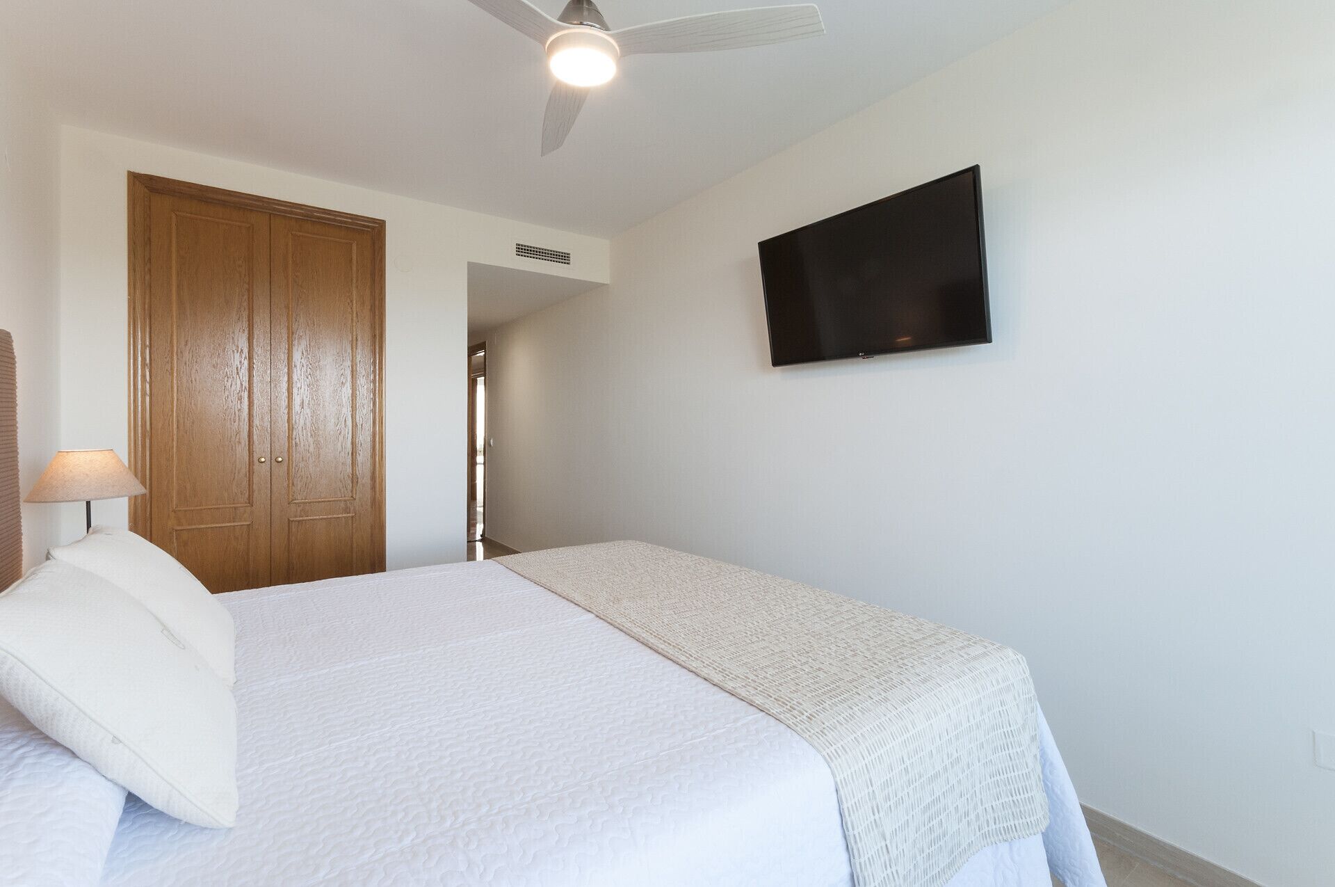 Ein Schlafzimmer mit Bett, Deckenventilator, Fernseher an der Wand und Holztür.