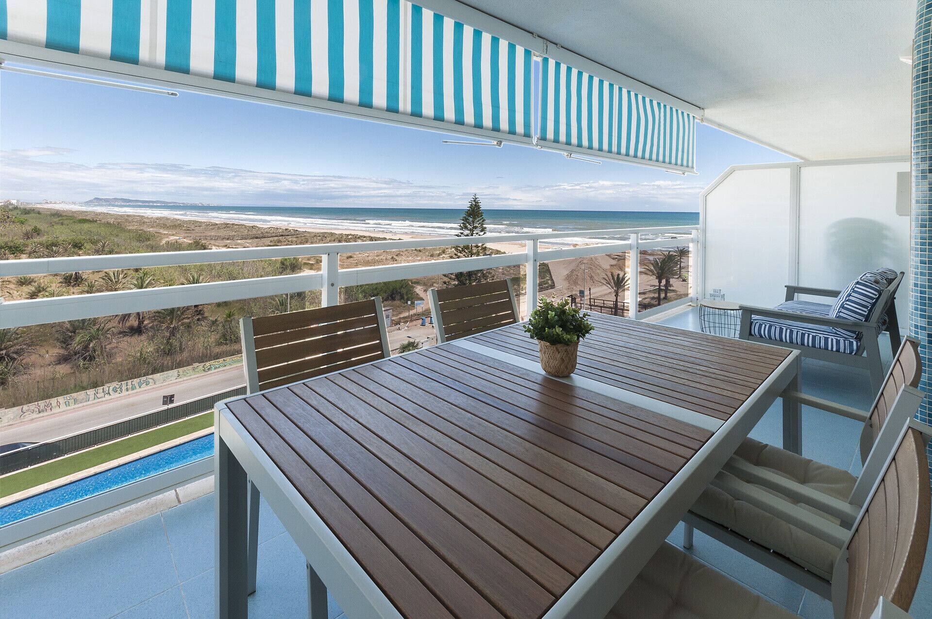 A balcony with a wooden table, a potted plant, and a view of the beach and ocean.