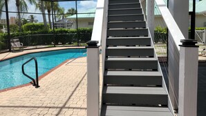 Un área de piscina con una escalera que conduce a una terraza cubierta, rodeada de palmeras y un cielo despejado.