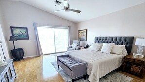 Un dormitorio con una cama grande, un banco tapizado en gris, una mesita de noche, una lámpara, un cuadro enmarcado y una ventana con cortinas.