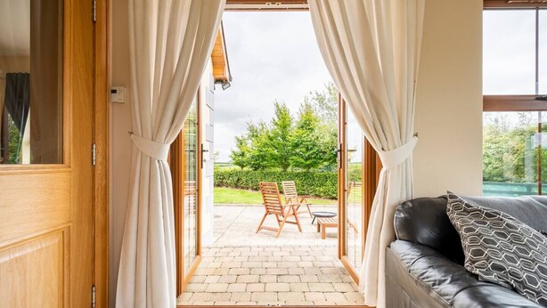 Et værelse med en trædør, der fører til en udendørs terrasse med et bord og stole, en sort lædersofa og udsigt til grønne områder.