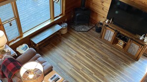 Ein gemütliches Blockhaus-Interieur mit einer Couch, Fernseher, Kamin und großen Fenstern mit Blick auf eine Terrasse.