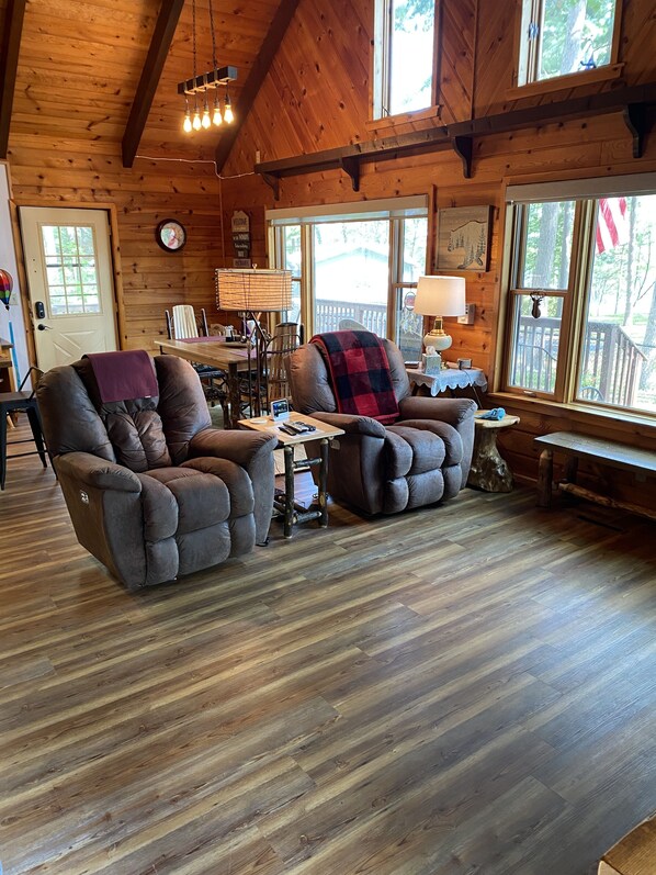 A cozy cabin interior with wooden walls and floors, two recliner chairs, a dining table, and a window with a view of a wooden deck.