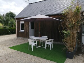 A wooden building with a gravel driveway, a white table and chairs under a large umbrella, and a potted plant.