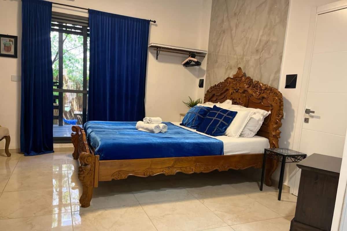 A bedroom with a wooden bed, blue bedding, and a view of the outdoors through a window with blue curtains.