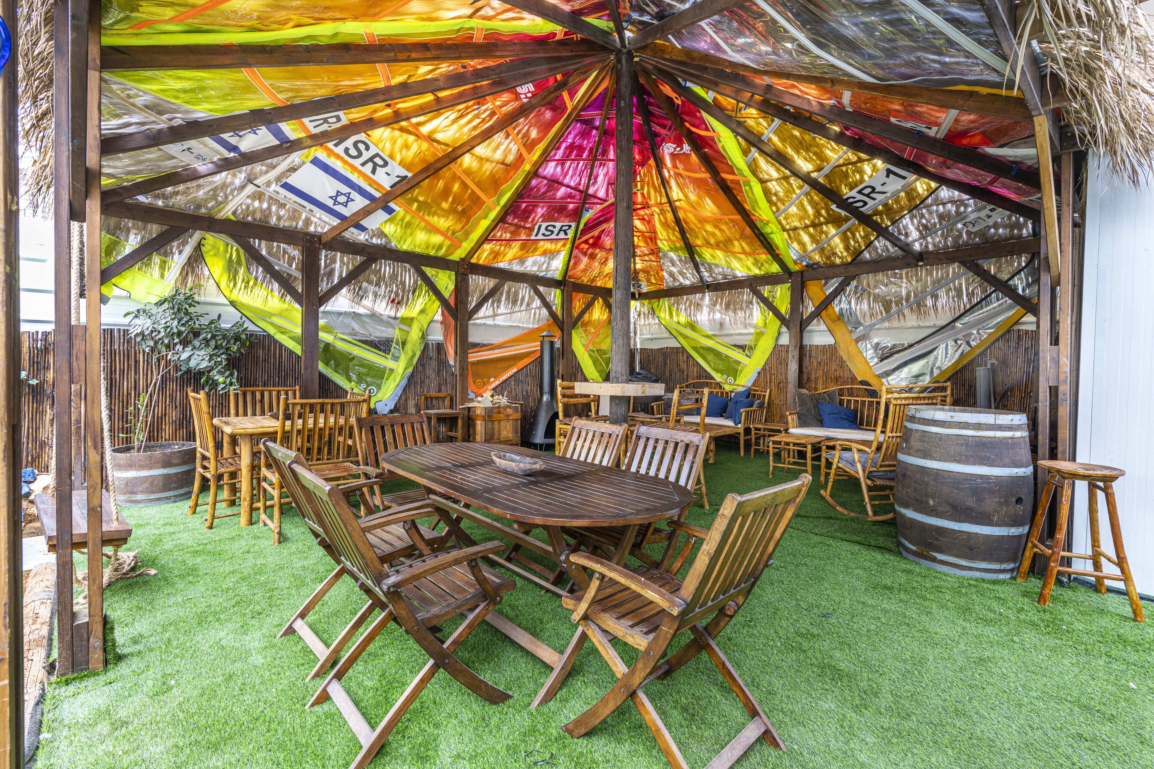 A covered outdoor seating area with a wooden table and chairs, surrounded by barrels and a thatched roof.