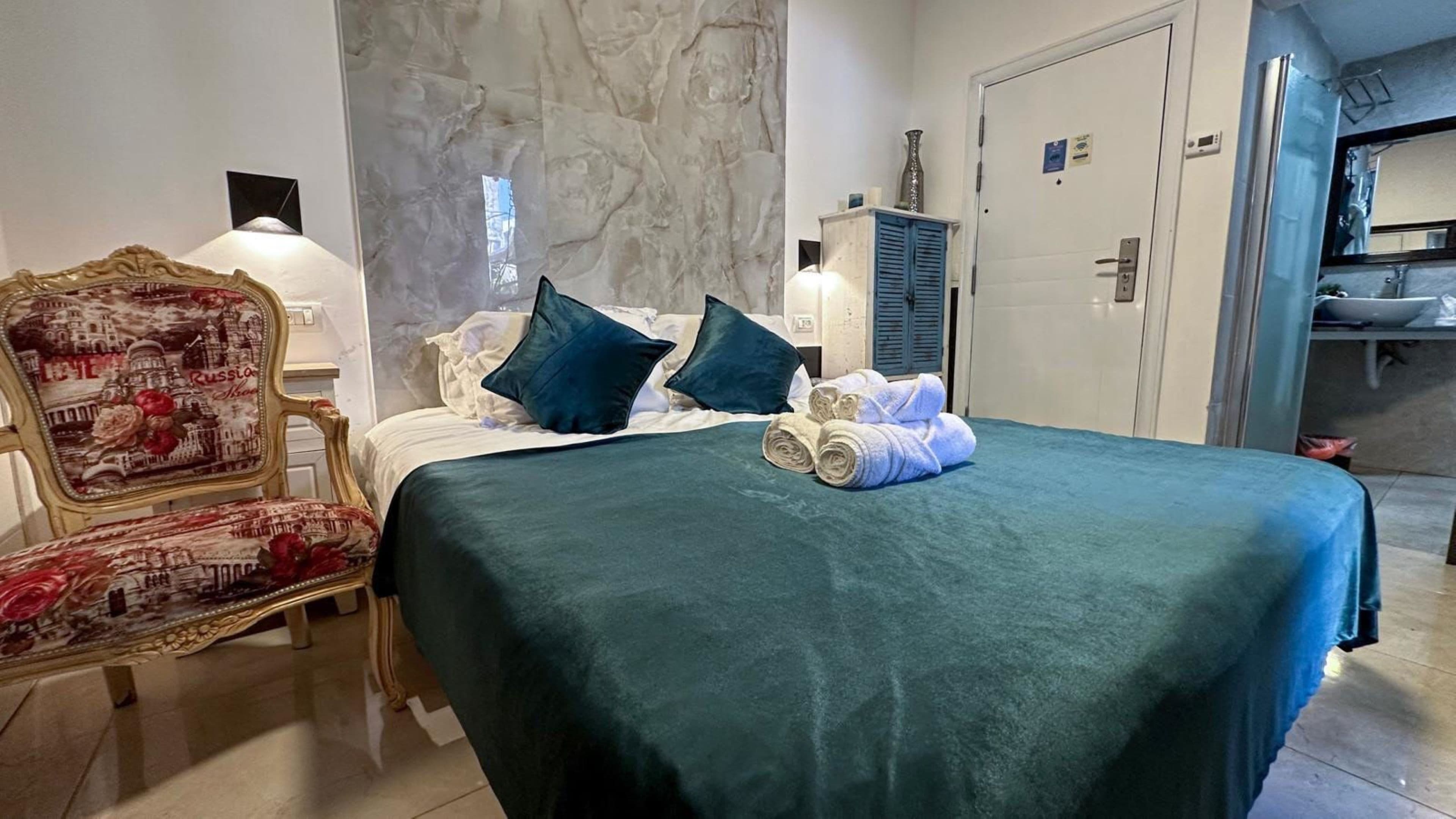 Una cama bien hecha con colcha color verde aguamarina, dos almohadas azul oscuro y un par de toallas enrolladas. Una silla de estilo vintage con tapicería floral se encuentra a la izquierda de la cama.
