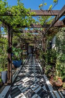 Un sendero cubierto con piso a cuadros, rodeado de diversas plantas y una estructura de pérgola de madera.