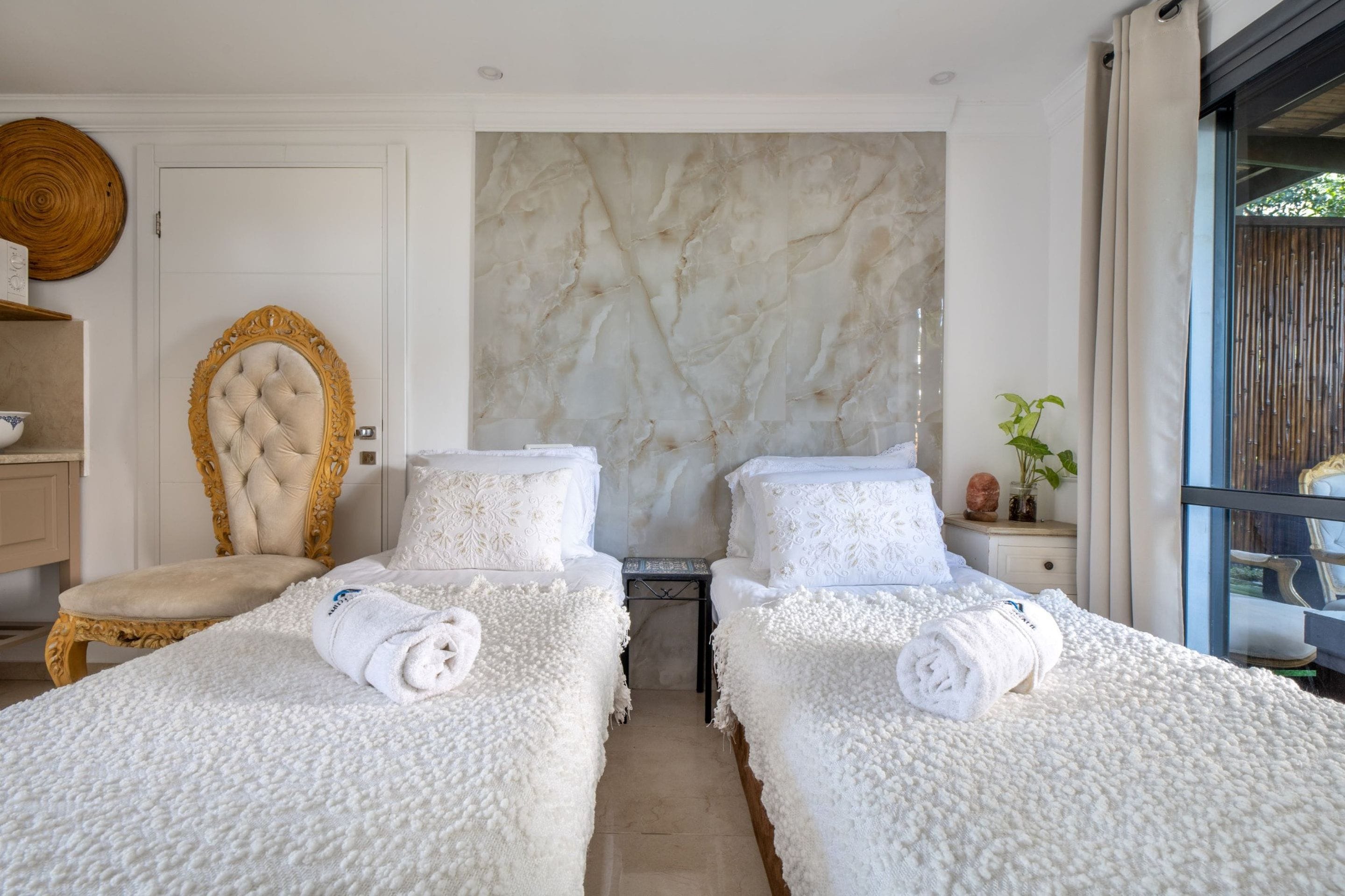 Habitación de hotel con dos camas, pared de mármol, una silla de madera y una mesita con una planta.