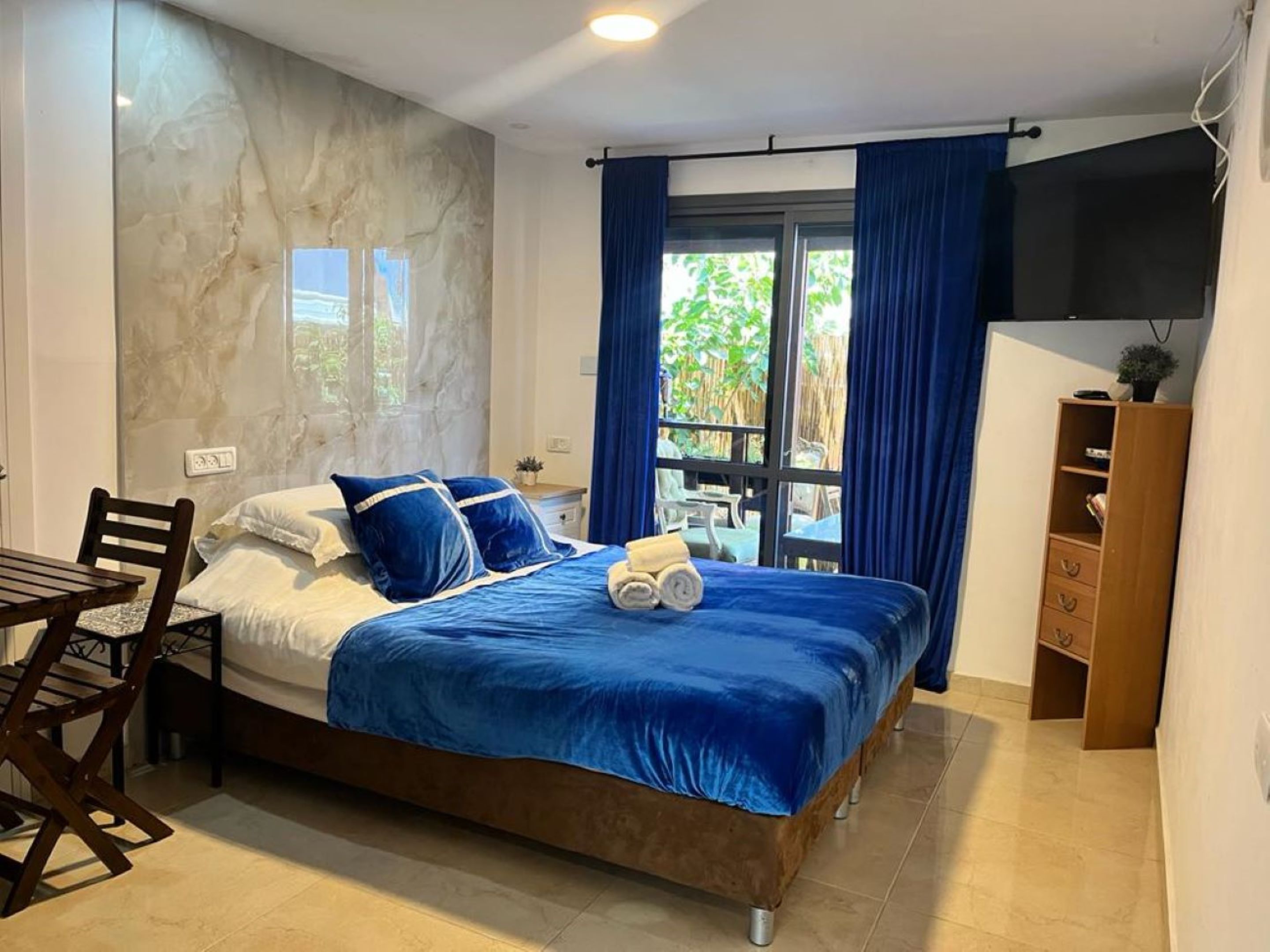 A bedroom with a bed, a wooden chair, a small table, a TV, and a window with curtains.
