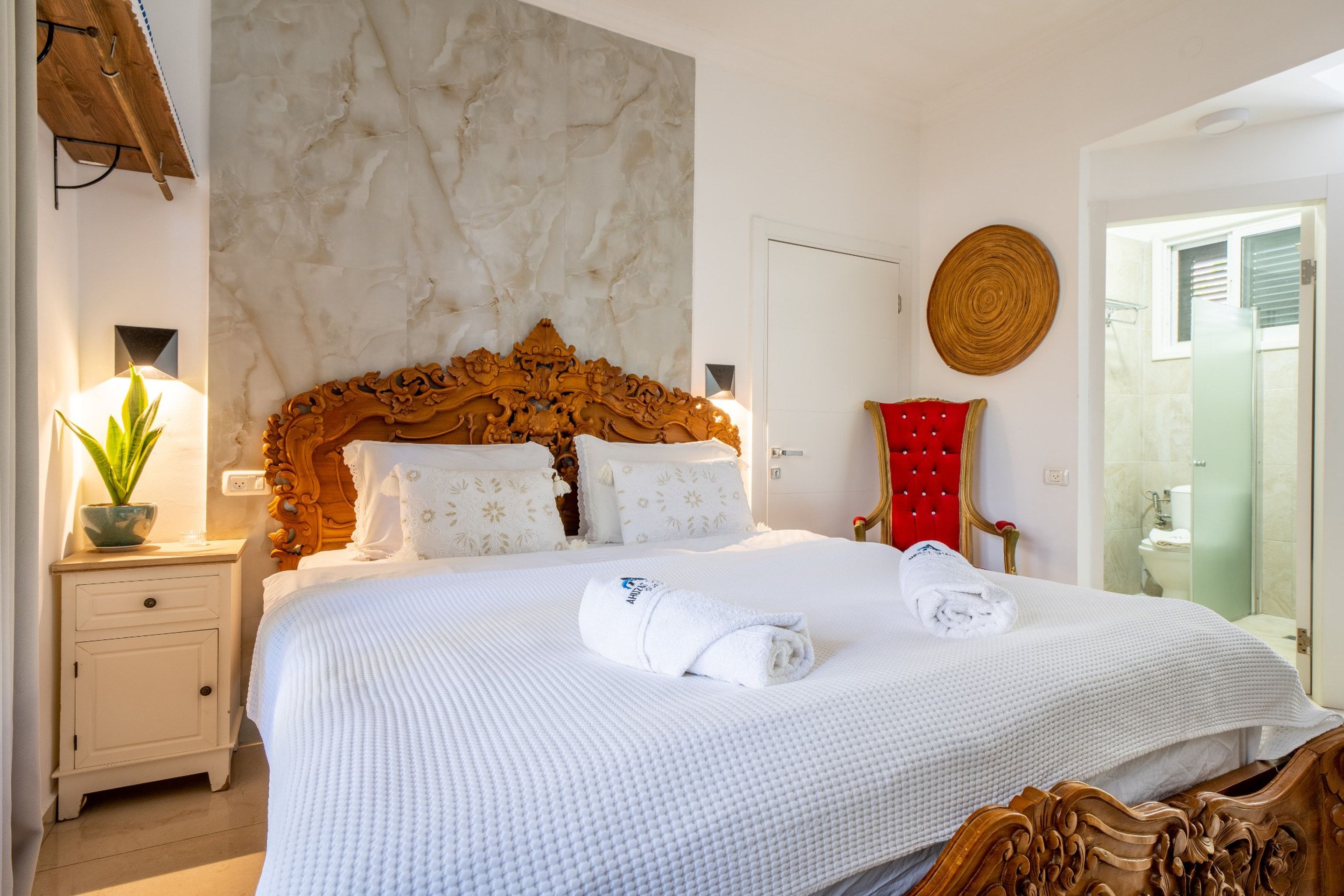 A hotel room with a marble wall, a wooden headboard, a red chair, a glass shower, and a white bed with towels.